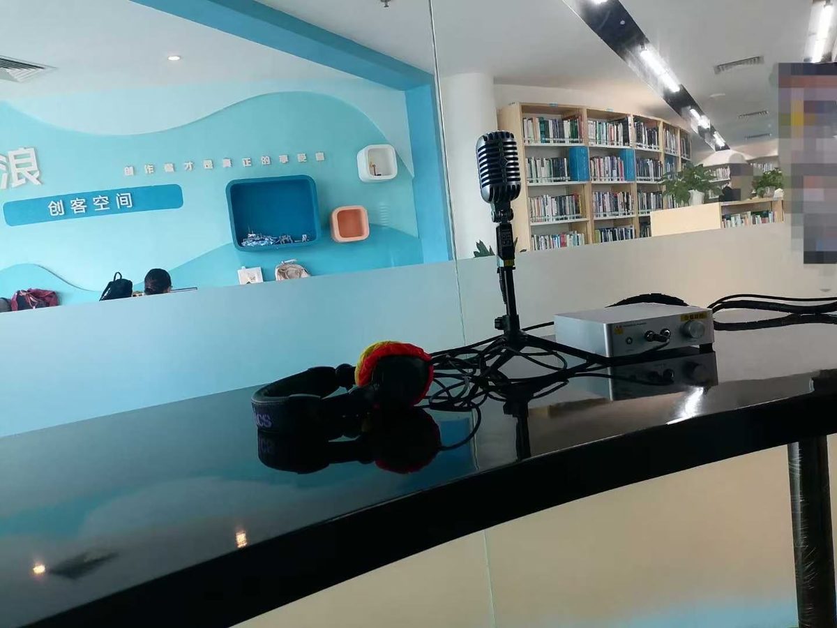 盐田区图书馆"创客空间"角落的一张设备照：墙上贴蓝色浪花海报与"创客空间"字样，桌面上摆着一支竖立的银色麦克风、一副红色的耳机和一台小型音频接口。背景右侧能看到图书馆书架
